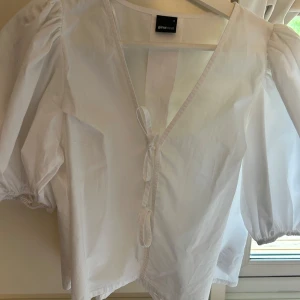Vit blus med puffärm från Gina Tricot - Säljer en vit blus från Gina Tricot med puffiga korta ärmar och v-ringning. Blusen har knytning framtill och är gjord i bomull, vilket gör den både luftig och snygg.