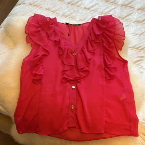 Rosa blus med volanger och guldknappar - Supersöt rosa blus med volanger längs v-ringningen och axlarna. Blusen har guldiga knappar framtill och är ärmlös, perfekt för en färgstark look. Tunt, lätt material som ger ett luftigt intryck och snyggt fall.
