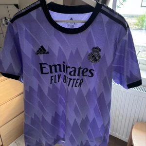 Lila Real Madrid fotbollströja Adidas - Snygg lila Real Madrid fotbollströja från Adidas med svart logga och klubbmärke på bröstet. Tröjan har ett geometriskt mönster, svarta detaljer vid krage och ärmslut samt trycket 'Emirates Fly Better' på framsidan. Tillverkad i lätt och ventilerande material.