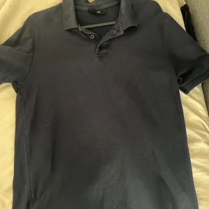 Mörkblå pikétröja från SMOG - Snygg mörkblå pikétröja från SMOG med klassisk krage och knappar framtill. Regular fit-modell som passar perfekt till jeans eller shorts. Tröjan har korta ärmar och är tillverkad i mjuk bomull för en skön känsla hela dagen.