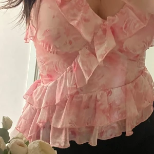 Rosa volangtopp med blommigt mönster - Supersöt rosa topp med volanger och blommigt mönster. Toppen har v-ringning och flera lager volanger upptill och nertill. Som ny, inte använt. 