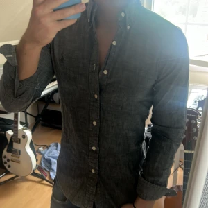 Mörkgrå skjorta från Ralph Lauren - Säljer en mörkgrå skjorta från Ralph Lauren med klassisk broderad logga på bröstet. Skjortan har lång ärm, knappar hela vägen och button-down krage. Perfekt till jeans för en clean look.