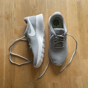 Gråa Nike sneakers med vit sula - Säljer ett par stilrena gråa Nike sneakers med vit sula och klassisk vit swoosh på sidan. Skorna har snörning och är tillverkade i syntetmaterial med meshstruktur för bra andningsförmåga. Perfekta för vardagsbruk eller lätt träning.