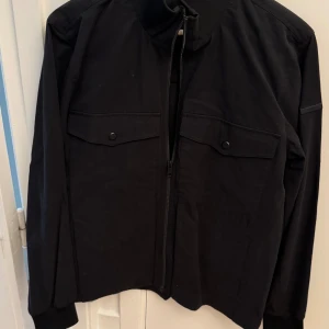 Svart jacka från Elvine med dragkedja - Snygg svart jacka från Elvine i storlek M. Jackan har dragkedja framtill, två bröstfickor med lock och knappar samt ribbad krage. Materialet är en mix av bomull och syntet, vilket gör den både bekväm och stilren.