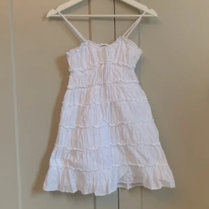 Vit somrig klänning från Gina Tricot - Supersöt vit klänning från Gina Tricot med smala axelband och volangkant nertill. Klänningen har broderade detaljer och knyt framtill, perfekt nu i sommar! Säljer då den tyvärr inte längre passar💕