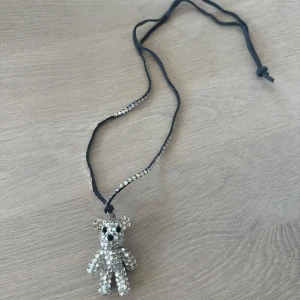 Halsband med glittrig björnberlock - Säljer ett halsband med svart band och en stor glittrig björnberlock täckt av silvriga stenar. Bandet har även silvriga detaljer längs ena sidan. Perfekt för dig som gillar unika accessoarer med lite extra bling.