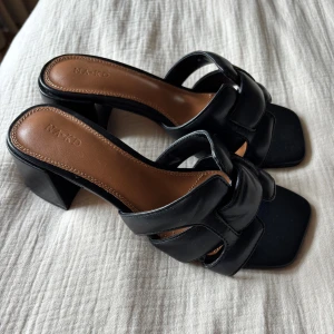 Svarta klackade sandaler från NA-KD - Säljer ett par svarta sandaler från NA-KD med breda remmar och fyrkantig tå. Skorna har en hög blockklack och är tillverkade i ett glansigt material med brun innersula. Perfekta för att lyfta vilken outfit som helst. Aldrig använda