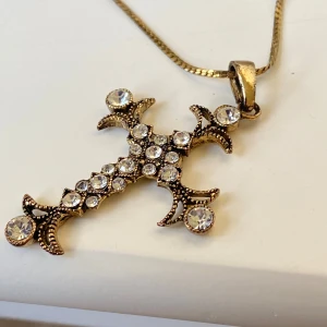 Guldfärgat halsband med kors och stenar - Säljer ett guldfärgat halsband med ett stort kors som hänge. Korset är dekorerat med gnistrande stenar och har en vintageinspirerad design. Perfekt statement-piece för dig som gillar unika smycken.