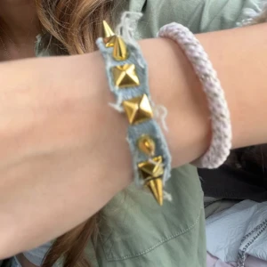 Armband med nitar och denim - Coolt armband i ljusblå denim med stora guldiga nitar i både kon- och pyramidform. Perfekt accessoar för att ge din outfit edge och attityd.