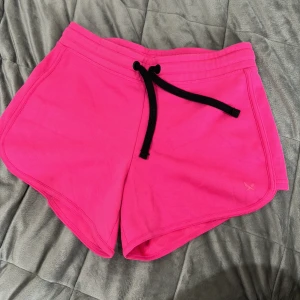 Rosa träningsshorts från SOC - Säljer ett par knallrosa träningsshorts från SOC i storlek XS. De har svart snörning i midjan och är gjorda i mjukt bomullsmaterial. Perfekta för gymmet eller varma dagar.