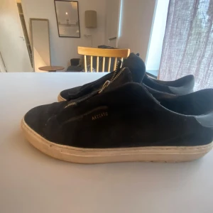 Svarta sneakers från Axel Arigato med dragkedja - Säljer ett par svarta sneakers från Axel Arigato med dragkedja framtill istället för klassisk snörning. Skorna har vit platt sula och är tillverkade i mocka med diskret logga på sidan.