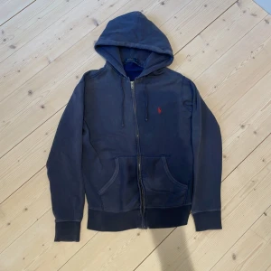 Mörkblå hoodie från Polo Ralph Lauren - Mörkblå hoodie från Polo Ralph Lauren med dragkedja och huva. Klassisk design med två fickor framtill och den ikoniska röda loggan broderad på bröstet. Skick 9/10, det är bara att skriva om ni undrar något!