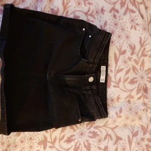 Svarta jeansshorts från You Denim - Svarta jeansshorts från You Denim med klassisk femficksdesign och knappgylf. Shortsen har bälteshällor och raka ben med uppvikta benslut. Perfekta för varma dagar och enkel att matcha med olika toppar.