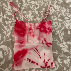 Rosa och vit topp med knytband - Säljer en snygg rosa och vit topp med batikmönster och tunna axelband. Toppen har omlottdesign med knytning i ryggen och croppad passform. Perfekt för dig som vill sticka ut lite extra!