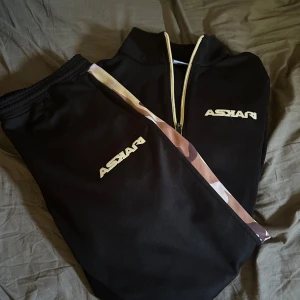 Svart träningsset från Askari med kamouflagedetaljer - Säljer ett svart träningsset från Askari med beige och bruna kamouflagedetaljer längs ärmar och byxben. Tröjan har dragkedja framtill och logga på bröstet, byxorna har resår i midjan och matchande logga.