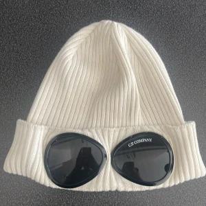 Vit ribbad mössa med goggles från C.P. Company - Säljer en vit ribbstickad mössa från C.P. Company med svarta inbyggda goggles framtill. Mössan har uppvikt kant och är tillverkad i mjukt material. Perfekt för dig som vill sticka ut med en unik accessoar.