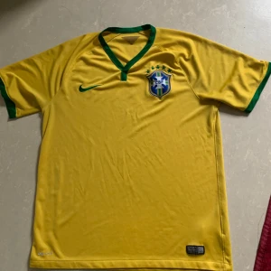 Retro Brasilien fotbollströja från Nike - Gul Brasilien fotbollströja från Nike med gröna detaljer vid krage och ärmslut. Tröjan har korta ärmar, broderad Nike-logga och Brasilien-emblem på bröstet. Tillverkad i lätt och ventilerande material, perfekt för match eller träning.