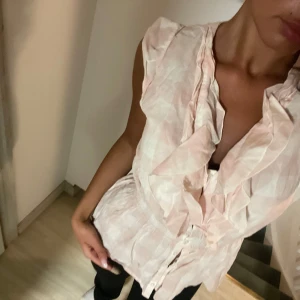 Ljusrosa blus med volang  - Säljer en ljusrosa blus med volangdetaljer längs den v-ringade halsen och knyt i midjan. Blusen är ärmlös och har en feminin, luftig design som passar perfekt till vår och sommar.