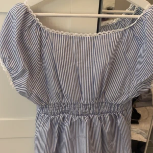 Blåvit randig blus med spetsdetaljer - Söt blåvit randig blus med korta puffärmar och smock i midjan. Blusen har fina spetsdetaljer längs halsringningen och ärmarna samt små knappar framtill. Perfekt för varma dagar.