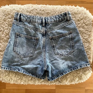 Blå jeansshorts från Lindex - Snygga blå jeansshorts från Lindex med fem fickor och bälteshällor. Modellen har normal passform och hög midja. Perfekta för sommaren och tillverkade i slitstarkt denimtyg. Kolla min profil för fler kläder!