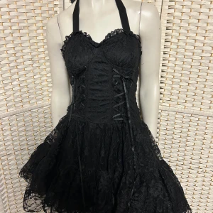 Klänning rockig storlek M fr Lucyfer - Supersnygg svart kortklänning med halterneck och vacker spets över hela klänningen. Livet har snörningsdetaljer framtill och en stor rosett baktill i midjan. Kjolen är voluminös och har volangkant. Perfekt för dig som vill sticka ut! Storlek M. Helt ny.