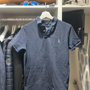 Mörkblå piké t-shirt från Ralph Lauren - Säljer en klassisk mörkblå piké t-shirt från Ralph Lauren med broderad logga på bröstet. T-shirten har krage, knappar och korta ärmar. Perfekt för en stilren och avslappnad look. Storlek medium men sitter som en small.