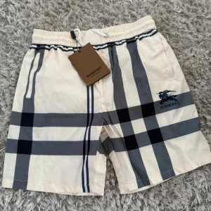 Burberry rutig shorts med snörning - Snygga Burberry shorts i klassiskt beige med stora svarta och grå rutor. Shortsen har elastisk midja med vit snörning och sidofickor. Tillverkade i Italien och märkta med Burberry-logga på benet. Perfekta för sommaren.