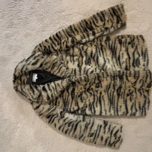Fuskpälsjacka med tigerprint från NA-KD - Säljer en snygg fuskpälsjacka från NA-KD med tigerinspirerat mönster i beige och svart. Jackan har lång ärm, krage och är fodrad på insidan. Perfekt statement-plagg för dig som vill sticka ut.