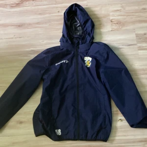 Blå vindjacka från Craft IFK Göteborg - Mörkblå vindjacka från Craft med IFK Göteborgs klubbmärke på bröstet. Jackan har huva, dragkedja framtill och långa ärmar. Perfekt för träning eller utomhusaktiviteter. Juniorstorlek 158/164. och Jackan är i perfekt skick