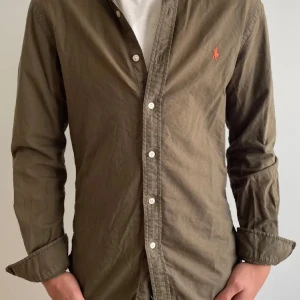 Olivgrön skjorta från Ralph Lauren - Säljer en olivgrön skjorta från Ralph Lauren med klassisk krage och knappar framtill. Skjortan har lång ärm och den ikoniska broderade loggan i orange på bröstet. Perfekt för dig som gillar stilrena plagg med snygga detaljer. För liten för mig derfor jag säljer