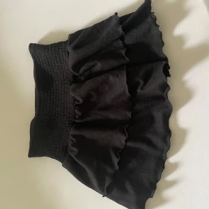 Svart volangkjol från Gina Tricot - Supersöt svart kjol från Gina Tricot med bred smock i midjan och dubbla volanger. Kjolen är kort och har en mjuk, stretchig känsla som gör den bekväm att bära. Perfekt att styla med en enkel topp eller hoodie.