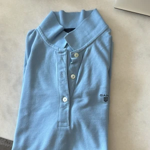 Ljusblå pikétröja från Gant - Säljer en klassisk ljusblå pikétröja från Gant med krage och knappar framtill. Tröjan har korta ärmar och diskret logga på bröstet. Perfekt för en stilren och avslappnad look.