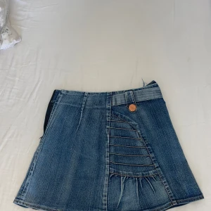 Jeanskjol med unika sömdetaljer - Säljer en cool jeanskjol i klassisk blå denim med dekorativa sömmar och knappar framtill. Kjolen har en kort passform och markerad midja med bälteshällor. Perfekt för dig som gillar en trendig och avslappnad stil.