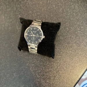 Silverfärgad klocka från Regal med svart urtavla - Stilren klocka från Regal med svart urtavla och silverfärgat armband i metall. Urtavlan har flera små visare och tydliga markeringar. Passar dig som gillar klassisk design och metallarmband.