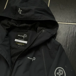 Svart regnjacka - Säljer en svart regnjacka från Pelle P med huva. Jackan har logotyp på bröstet och ärmen samt justerbar huva. Perfekt för båtliv, blåsiga och regniga dagar. Nypris 2800kr, Storlek S men sitter M