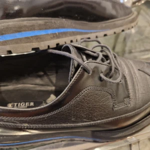 Svarta skinnloafers från Tiger of Sweden - Säljer ett par svarta loafers i skinn från Tiger of Sweden. Skorna har snörning, rund tå och en svart sula med blå detalj. Perfekta för dig som gillar stilrena och klassiska skor med en modern twist.