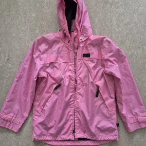Vindjacka - Säljer en rosa vindjacka från Active mywear i storlek 140. Jackan har huva, dragkedja framtill och två snedställda fickor med dragkedja. Finns några fläckar. Perfekt för blåsiga dagar och enkel att ta med sig. Snygg och praktisk design.