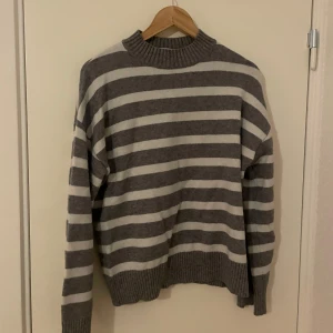 Gråvit randig stickad tröja från Kappahl - Mysig stickad tröja från Kappahl i grått och vitt med breda horisontella ränder. Tröjan har rund halsringning, långa ärmar och ribbade muddar vid ärmslut och nederkant. Perfekt för kyligare dagar.