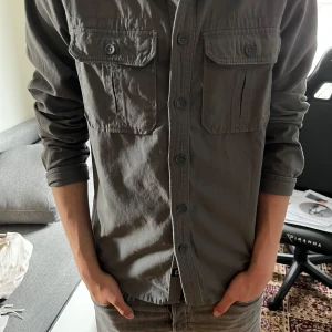 Mörkgrå overshirt  - Säljer en stilren mörkgrå overshirt med två stora bröstfickor och knappar framtill. Overshirten är från Brandit, modellen på bilden är 183cm lång. Vid fråger eller funderingar är det bara att höra av sig. 