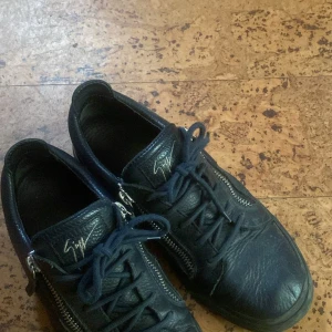 Svarta sneakers med dragkedja från Giuseppe Zanotti - Säljer ett par svarta sneakers från Giuseppe Zanotti i skinn med snörning och dragkedjor på sidorna. Skorna har rund tå och platt sula, samt den ikoniska signaturen på plösen. Tillverkade i Italien.