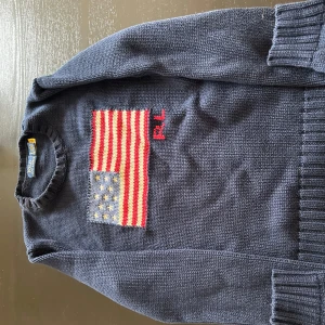 Mörkblå stickad tröja med amerikansk flagga från Ralph Lauren - Säljer en mörkblå stickad tröja från Polo Ralph Lauren med en stor amerikansk flagga framtill och RL-broderi i rött.Tröjan är i perfekt skick bara används en gång. Tröjan har rund halsringning och långa ärmar. Perfekt för dig som gillar cleangirl stilen, old money, stocholmstil eller något annat . Säljer den för 2000 altså 50% billigare än om du skulle köpa en ny (4000 är orginal pris)