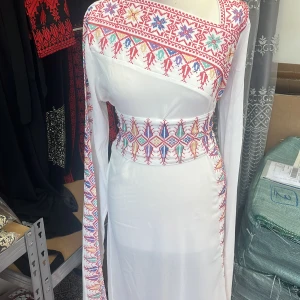 Vit långklänning med broderade detaljer - Säljer en vit långklänning med färgglada broderade mönster i rött, blått och rosa längs ena axeln, midjan och ärmarna. Klänningen har en omlottliknande axel och knytband i midjan för en markerad siluett. Perfekt för dig som gillar unika och traditionella detaljer.