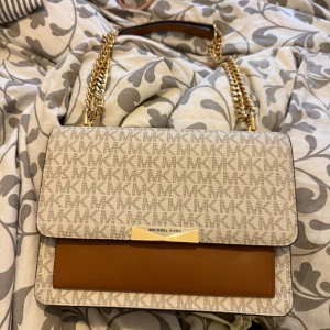 Michael Kors axelväska med kedjerem - Snygg axelväska från Michael Kors i beige och brunt med MK-logga över hela väskan. Väskan har guldfärgade kedjeremmar och detaljer samt flera fack inuti, inklusive dragkedja. Perfekt för att lyfta din outfit.