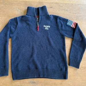 Marinblå stickad tröja med halv dragkedja från GANT - Mörkblå stickad tröja från GANT med halv dragkedja och hög krage. Amerikansk flagga på ena ärmen och GANT Est. 1949-tryck på bröstet. Tillverkad i 100% bomull