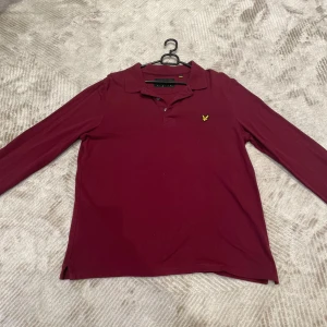 Vinröd pikétröja från Lyle & Scott - Säljer en vinröd långärmad pikétröja från Lyle & Scott med klassisk krage och knäppning framtill. Tröjan har det ikoniska gula fågel-loggan broderad på bröstet. Perfekt för en stilren och avslappnad look.