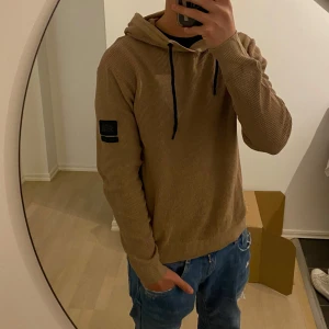 Beige hoodie med huva och svarta snören - Säljer en beige hoodie från Jack & Jones med huva och svarta snören. Tröjan har lång ärm, ribbade muddar och en patch på vänster ärm. Perfekt för en avslappnad stil.
