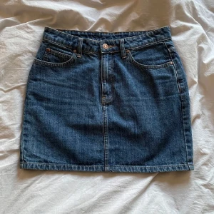 Jeanskjol från Dr Denim - Jättefin mörkblå jeanskjol från Dr Denim med klassisk femficksdesign och knappgylf. Kjolen har bälteshällor och raka sömmar, perfekt för en avslappnad stil. Enkel och tidlös modell som passar till mycket. 