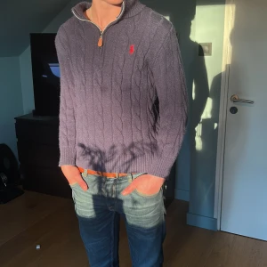 Marinblå  stickad tröja med dragkedja från Ralph Lauren - Säljer en lila stickad tröja från Ralph Lauren med halv dragkedja och röd broderad logga på bröstet. Tröjan har kabelstickat mönster och hög krage. Perfekt för kyligare dagar. Mannen på bilden är 175 cm lång