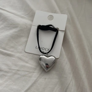 Halsband med silverfärgat hjärta från Lindex - Snyggt halsband från Lindex med ett stort, blankt hjärta i silverfärg. Hängs i ett svart band som ger en cool kontrast till det glansiga hjärtat. Perfekt accessoar för att lyfta din outfit. Nypris är 60kr så tänker jag tar 30kr för den😁😁