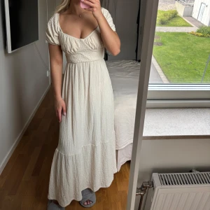 Vit långklänning med puffärmar och knyt i ryggen - Superfin vit långklänning med puffärmar och markerad midja. Klänningen har v-ringad urringning och snygg knytning i ryggen. Perfekt för sommarens alla tillfällen. Materialet känns lätt och luftigt. Aldrig använd 